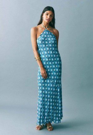 Rochie maxi din țesătură teal, cu un model geometric alb, cu decolteu halter și o ușoară evazare la tiv, asortată cu accesorii aurii.