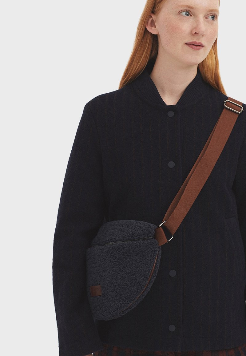 Borsa a tracolla in pile grigio scuro con forma curva, cinghia marrone e chiusura con zip; presenta una piccola toppa decorativa. Indossata sopra un cappotto a righe nere.
