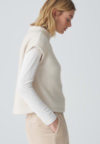 Blonde vrouw in een beige gewatteerde mouwloze vest over een witte langslipshemd, met haar handen in de zakken van een beige corduroy broek, in zijaanzicht.