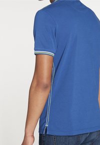 Blå pikétröja med korta ärmar, med vita och gröna randiga detaljer på kragen och ärmarna, samt slitsar i sidorna vid fållen.