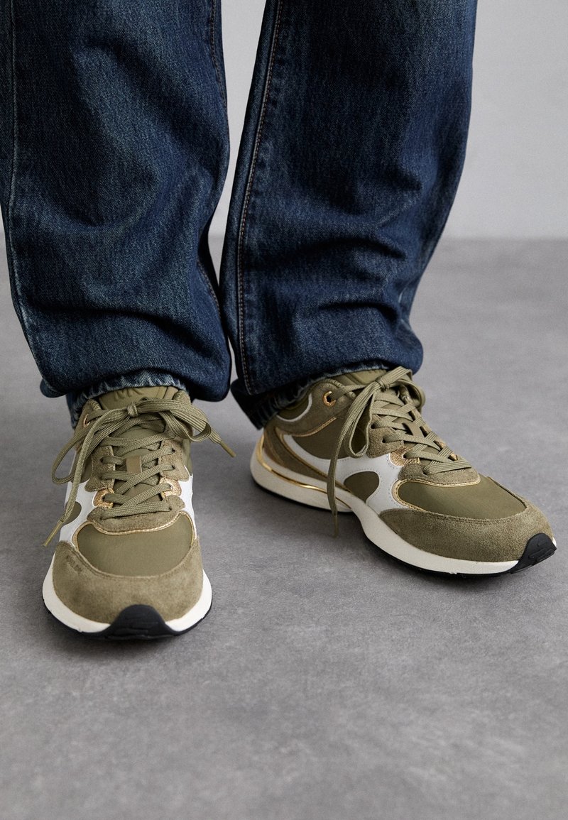 Personne portant des baskets vertes et blanches avec des semelles blanches et un jean bleu foncé, debout sur un sol gris.