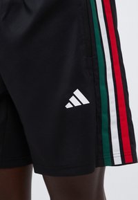 Μαύρο αθλητικό σορτς με κατακόρυφες ρίγες σε πράσινο, λευκό και κόκκινο στο πλάι και λευκό λογότυπο Adidas κοντά στο τελείωμα.