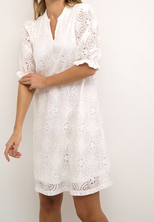Woman wearing a knee-length white lace dress with floral patterns and puffed sleeves, adjusting the left sleeve with her right hand.