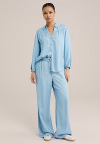 Hellblaues Chambray-Hemd mit Knöpfen, lockerer Passform, langen Ärmeln und gerüschtem Detail. Kombiniert mit weit geschnittenen passenden Hosen und Slip-On-Schuhen.