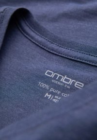 Modrá bavlněná tkanina trička s názvem značky "Ombre"; obsahuje štítek s péčí uváděj�ící "100% čistá bavlna" a velikost "M" na vnitřním límci.