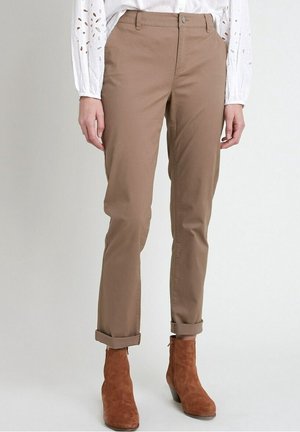 Vrouw draagt beige opgerolde broek, bruine suède enkellaarsjes en een witte blouse met lange mouwen en oogjesdetails.