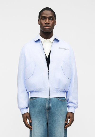 Hombre con chaqueta azul claro con cremallera sobre una camiseta blanca y chaleco negro, combinado con vaqueros azules, de pie frente a un fondo liso.