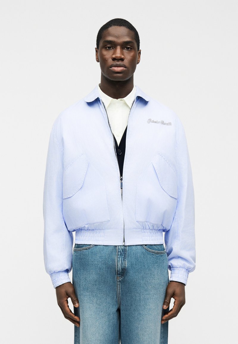 Homme portant une veste zippée bleu clair sur une chemise blanche et un gilet noir, associée à un jean bleu, debout devant un fond uni.