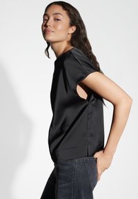 Schwarzes Satinoberteil mit kurzen Ärmeln und Ausschnitten an den Seiten, kombiniert mit dunklen Jeans. Glatte Textur und elegantes Design.