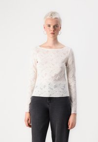 White lace long-sleeve top with floral pattern and a wide neckline, paired with dark denim jeans. Textured fabric with sheer areas.