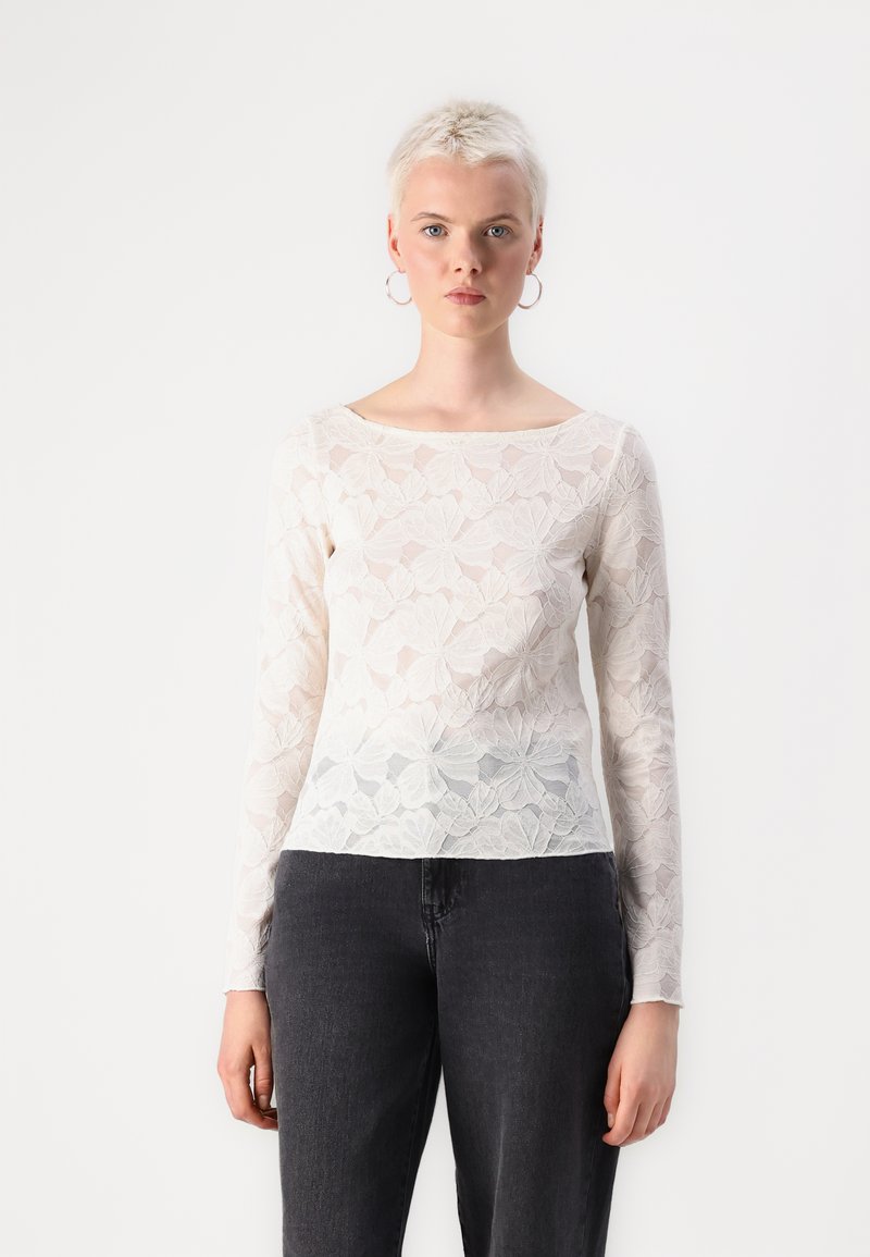 Hvit bluse med lange ermer laget av blonder og med blomsterdesign, kombinert med mørk denim jeans. Teksturiert stoff med gjennomsiktige områder.
