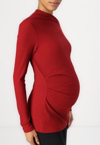 Raudona juostelėta ilgio rankovėm nėščiųjų palaidinė su prigludusiu siluetu, su šoniniu raukšlėjimu ir aukštu kaklu, derinama su juodomis legginsais.