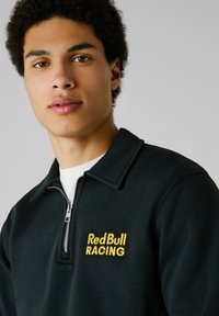 Zwarte kwart-rits sweatshirt met een platte kraag, geborduurd "RedBull RACING" logo in het geel, gemaakt van een zacht materiaal.