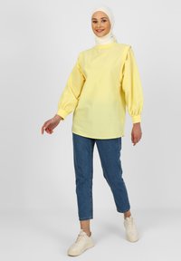 Haut à manches longues jaune avec un col haut et des manches bouffantes, associé avec un jean bleu et des baskets beiges. Texture de tissu lisse, coupe décontractée.