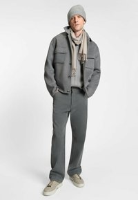 Mand stående iført grå hue, grå jakke, beige tørklæde, grå sweater, grå bukser og beige sneakers mod en ensfarvet hvid baggrund.