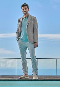 Light gray blazer over a light blue polo, paired with light gray trousers and beige shoes. Man stands on a deck beside a pool, with sky backdrop.