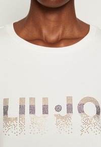 Zblízka osoba v bílém tričku s písmeny posetými zlatem a fialovou, která tvoří částečný text na hrudi.