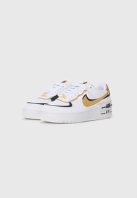 Białe i złote niskie sneakersy Nike Air Force z czarnymi detalami i grubymi podeszwami na jednolitym jasnoszarym tle.