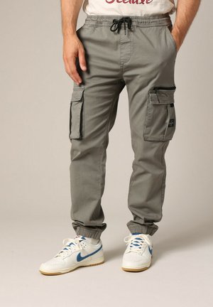 Pantalones cargo grises con puños elásticos, cintura con cordón, múltiples bolsillos, fabricados en un tejido ligero; combinados con zapatillas blancas con acentos azules.