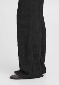 Schwarze, texturierte, weite Hosen mit einer gekräuselten Stoffoberfläche und seitlichen Schlitzen, kombiniert mit dunklen, spitzen Schuhen.