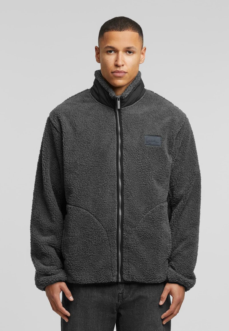 Fleece jacket in dark gray, featuring a high collar, zip closure, and textured fabric. Plain design with a side logo patch.