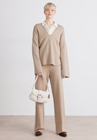 Beige knit sweater and matching pants, with a white collared shirt underneath. Holding a cream-colored handbag with silver hardware.