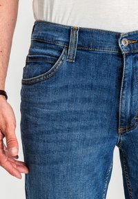 Niebieskie dżinsy wykonane z denimu o prostym kroju nogawek, z pięcioma kieszeniami i kontrastującym pomarańczowym przeszyciem wzdłuż szwów i kieszeni.