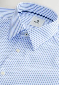Chemise à manches longues bleu clair et blanche à rayures, avec un design à boutonnière, un col et une étiquette en tissu portant le mot "NEXT". Des boutons sobres ornent le devant.