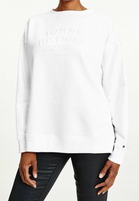 Persona che indossa una felpa bianca Tommy Hilfiger con testo in rilievo e pantaloni neri, in piedi contro uno sfondo neutro.