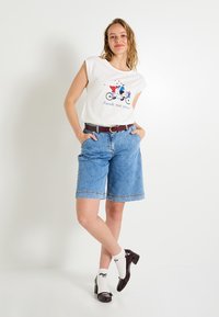 Camiseta blanca con un gráfico de bicicleta colorido, combinada con bermudas de mezclilla. Cinturón marrón, calcetines blancos con lazos negros y tacones oscuros de charol.