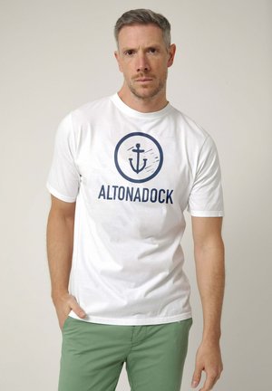 Hombre con cabello corto y canoso que lleva una camiseta blanca con un logo de ancla azul marino y el texto "ALTONADOCK", combinado con pantalones verdes, de pie frente a un fondo liso.