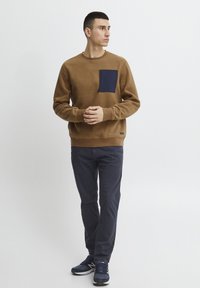 Sudadera de felpa marrón con un bolsillo en el pecho azul marino, cuello redondeado, puños y dobladillo acanalados, combinada con pantalones azul marino y zapatillas deportivas azul marino.