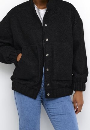 Personne portant une veste en laine noire boutonnée avec deux poches avant, des poignets élastiques, et un jean bleu clair, la main dans la poche gauche.