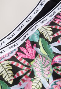 Tissu à imprimé floral coloré avec de grandes feuilles dans des nuances de rose, vert et noir. La ceinture blanche présente le logo "NEXT".