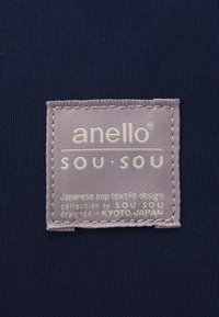Merkkitag sinivihreää kangasta, jossa on "anello" ja "sou-sou" -brändäys. Tagissa on teksturoitu harmaa tausta, valkoinen teksti ja beige ommel.