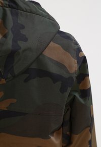 Camouflage-mønstret jakke med hætte i mørkegrønne og brune toner, lavet af glat, letvægtsstof med mat finish.