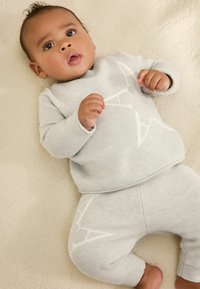 Heled halli kootud beebiriietus, millel on pika varrukaga ülemine osa, millel on geomeetriline valge mustriline disain ja sellele sobivad püksid, pehme beeži tekikese peal.