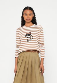 Žena v hnedom a bielo pruhovanom tričku s dlhým rukávom s grafickou potlačou kresleného mačky a plisovanou béžovou sukňou s opaskom, stojaca na bielom pozadí.