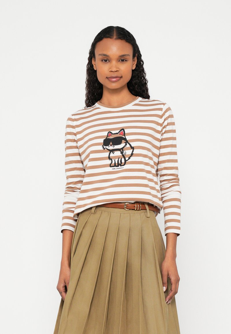 Žena v hnedom a bielo pruhovanom tričku s dlhým rukávom s grafickou potlačou kresleného mačky a plisovanou béžovou sukňou s opaskom, stojaca na bielom pozadí.