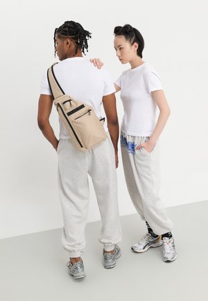 Beige crossbody bag med flere glidelåslommer, båret av en person i en hvit t-skjorte og grå joggebukser, satt mot en ensfarget hvit bakgrunn.