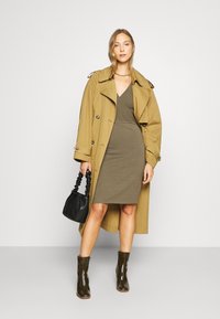 Trencho bege sobre um vestido ajustado verde-azeitona, combinado com botins brilhantes verde-azeitona e uma pequena bolsa preta texturizada.