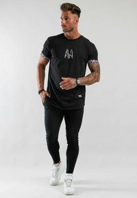 Svart bomullströja med kontrasterande blommiga ärmar och en logotyp, kombinerad med slim fit svarta byxor och vita sneakers. Diskreta texturerade detaljer.