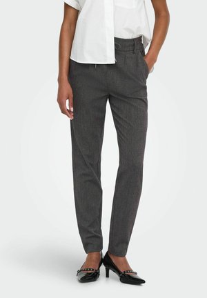 Femme portant un pantalon gris sur mesure, un chemisier blanc à manches courtes et des chaussures noires à talons moyens, debout avec une main dans la poche.