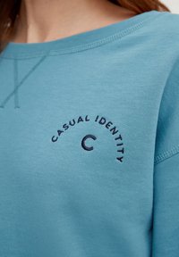 Sweatshirt turquoise avec col rond, arborant un texte brodé en bleu foncé "CASUAL IDENTITY" et une lettre "C". Texture de tissu lisse.