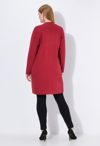 Rode lange mouwen jurk, gladde stof, halverwege de dij lengte, met zijslits, gecombineerd met zwarte leggings en hoge hakken.