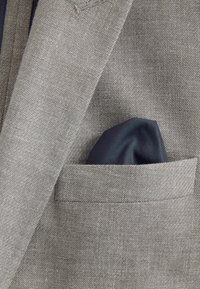 Veste de costume grise avec un mouchoir de poche bleu foncé soigneusement plié dans la poche poitrine.