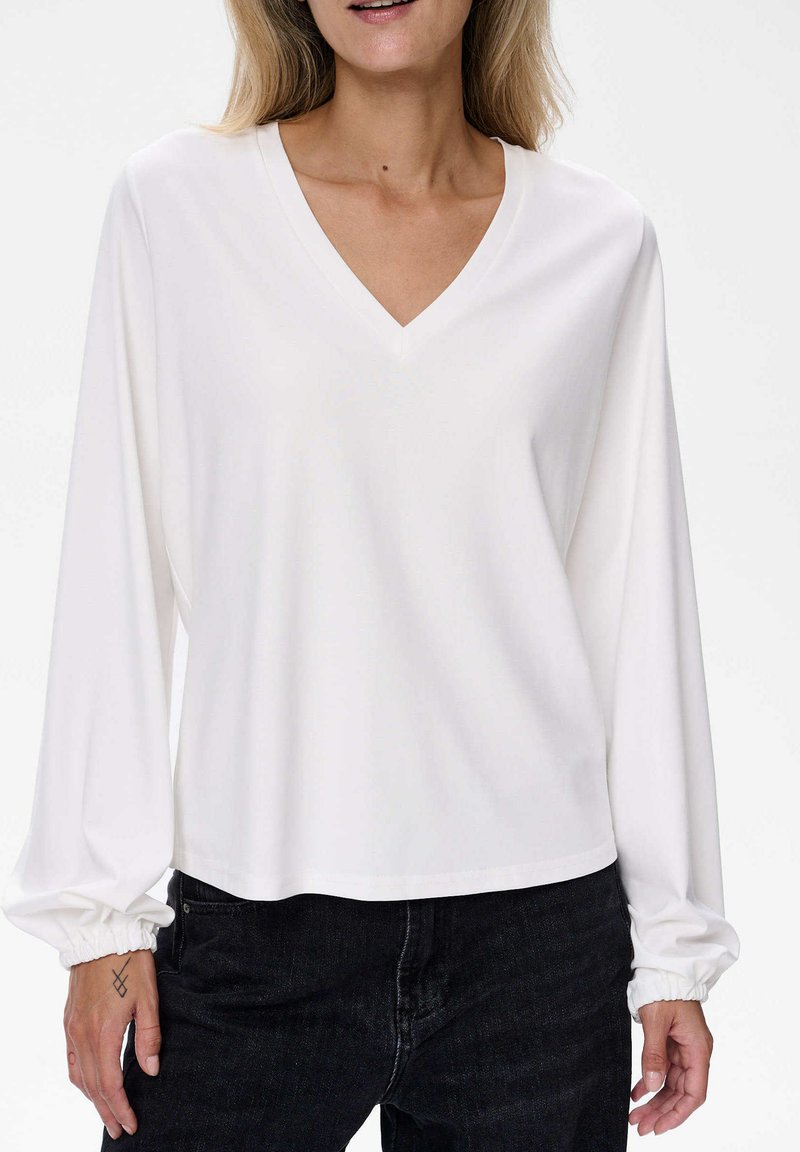 Weiße Langarm-V-Ausschnitt-Bluse mit elastischen Bündchen, aus glattem Stoff. Kombiniert mit dunklen Jeans. Einfaches und lockeres Design.