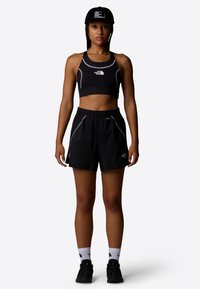 Top corto deportivo negro con logo blanco y detalles en azul, combinado con pantalones cortos negros con bolsillos laterales. El modelo lleva zapatillas negras y calcetines.