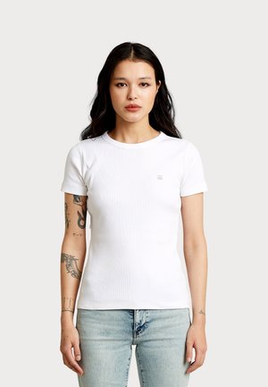 Vrouw met lang donker haar, die een eenvoudige witte korte mouwen T-shirt en lichtblauwe spijkerbroek draagt, en tatoeages op haar linkerarm laat zien.