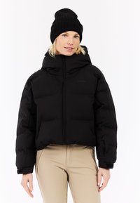 Čierna puffová bunda s vysokým golierom, so zipsom vpredu a bočnými vreckami. Nosená s čiernou pletenou čiapkou. Hladká textúra a skrátený dizajn.